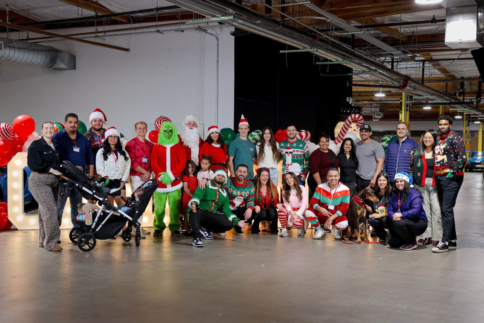 A big group of CARE volunteers and helpers in holiday outfits, with Santa, an elf and a green costumed character, smiling together at Claus for a Cause